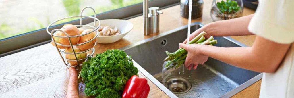 A woman washing asparagus in a sink with running water. There is potatoes, lettuce and other vegestables on the bench next to the woman who is wearing a white t-shirt